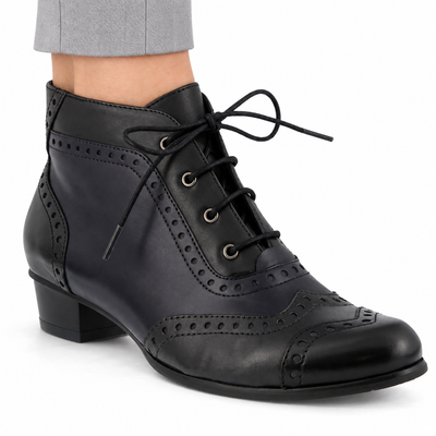 AI-generated on-model view of the Spring Step Heroic women's Black ankle boot with subtle dark navy panels, brogue detailing and black laces, worn with light gray trousers on a white background.